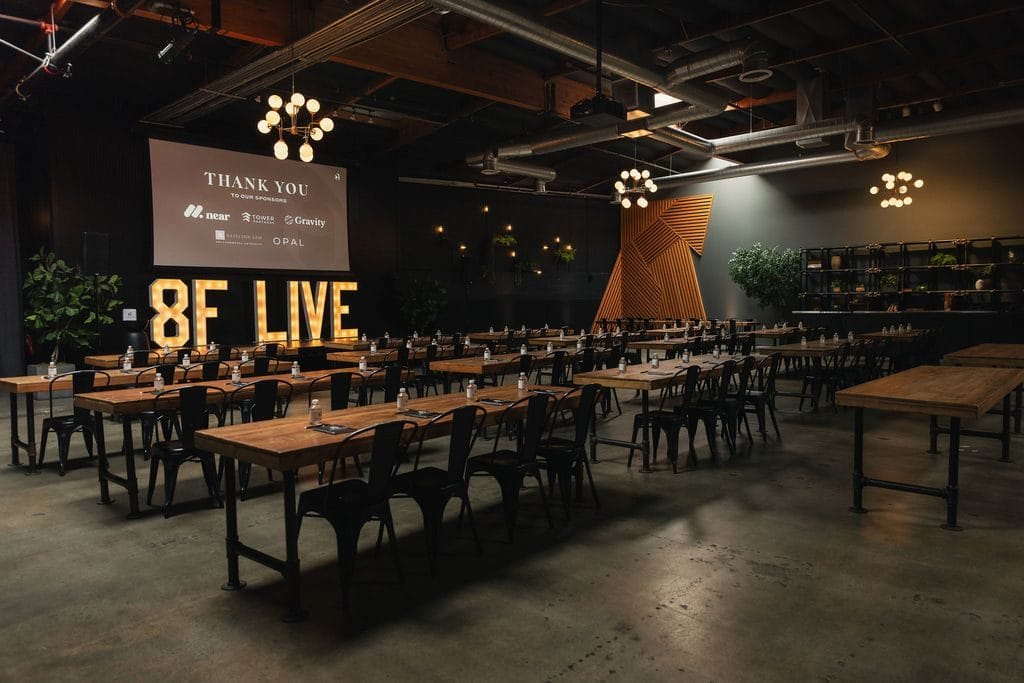 Long wooden communal tables facing illuminated 8F LIVE marquee letters and a sponsor screen