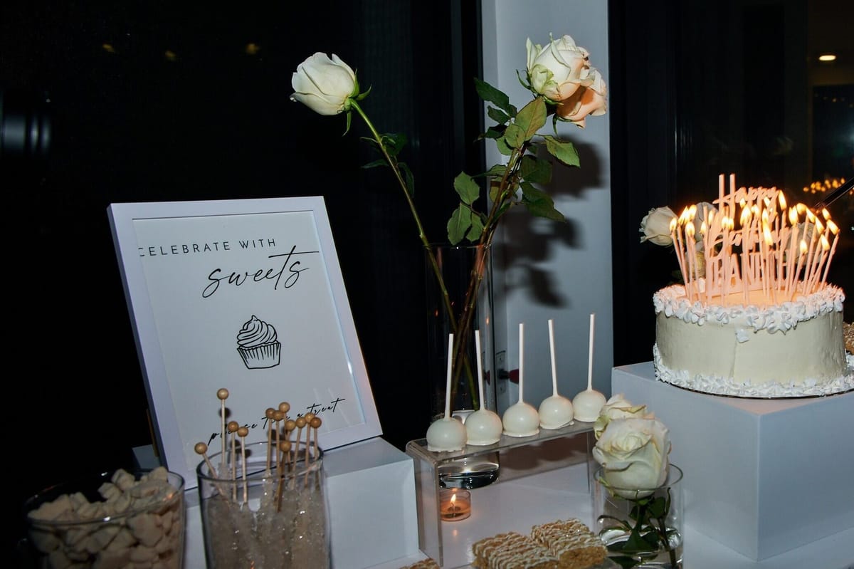 Sweets table at night with a Celebrate with sweets sign, white roses, white cake pops and a candlelit white cake