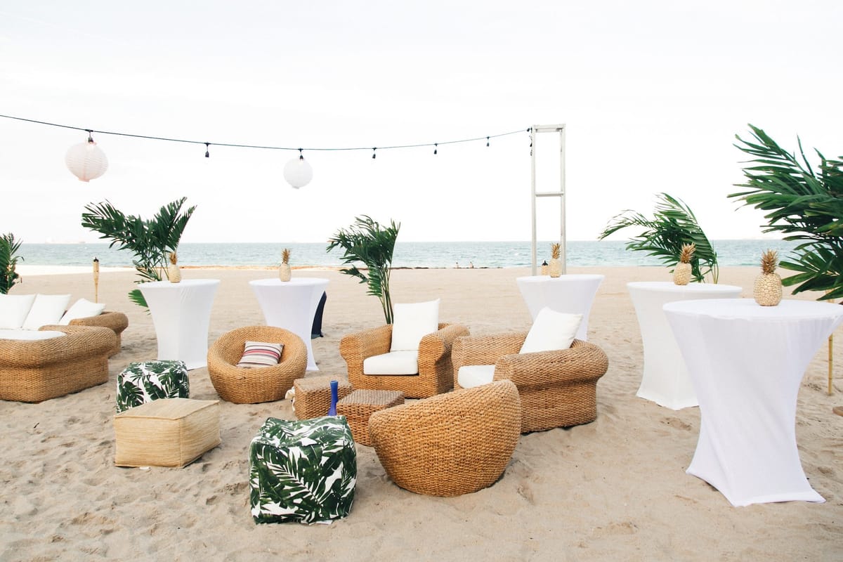 Wicker armchairs, palm-print poufs and white cocktail tables on the sand under string lights by the ocean