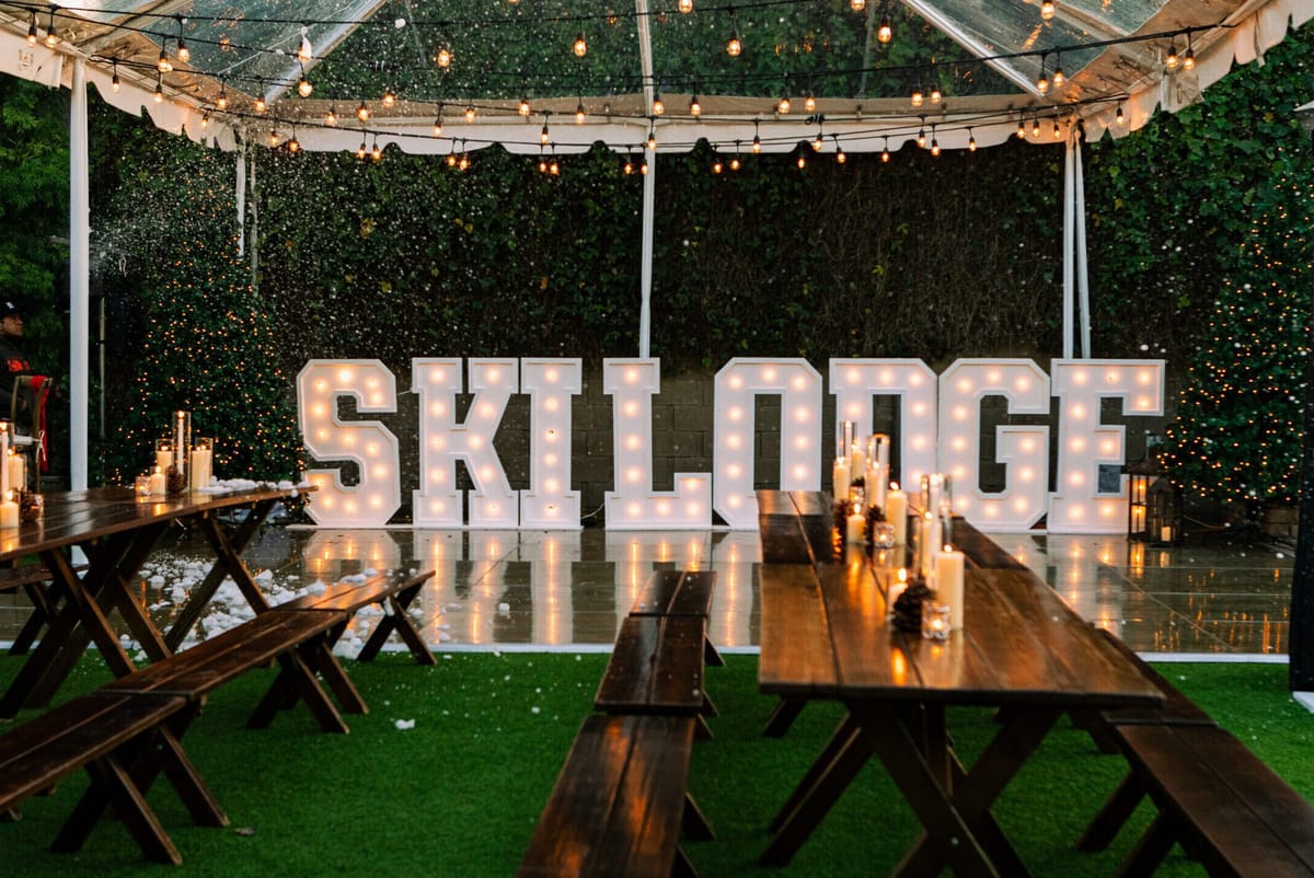 Clear tent with lit SKI LODGE marquee letters behind long wooden tables and benches