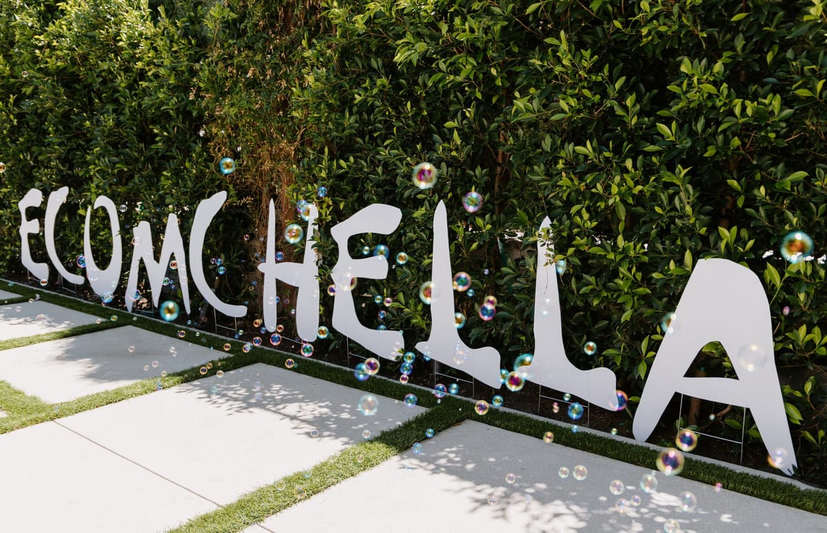Tall white ECOMCHELLA letters standing in front of a green hedge with soap bubbles floating past