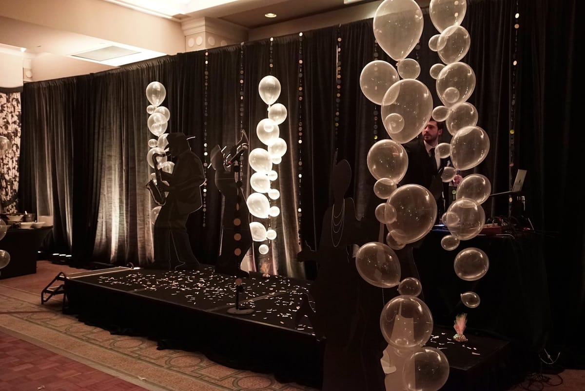 DJ stage with black drapes, clear balloon columns and black silhouettes of jazz musicians and a flapper, confetti on the floor