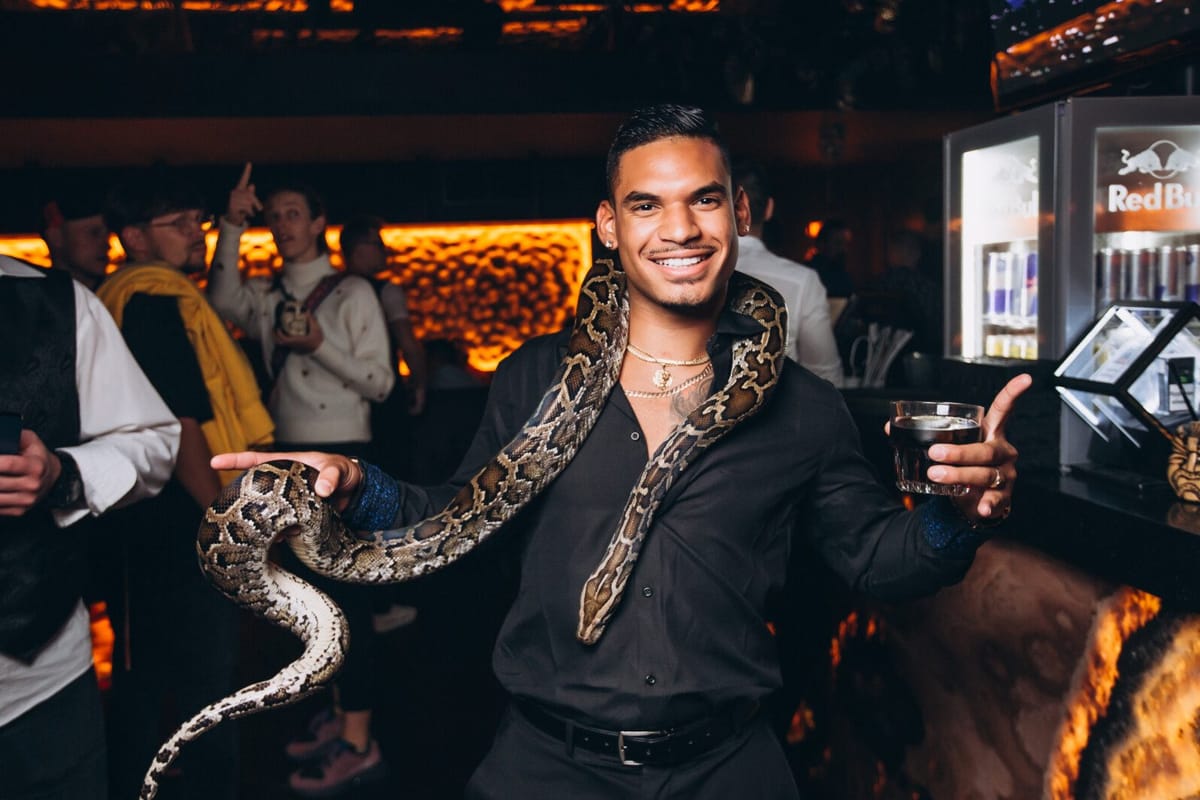 Smiling man holding a drink with a large snake draped around his shoulders in a dim room lit orange