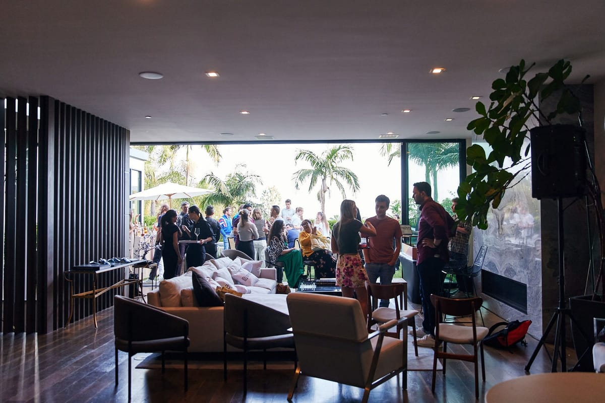Guests mingling in a living room with sofas and armchairs, glass walls open to palm trees and a terrace