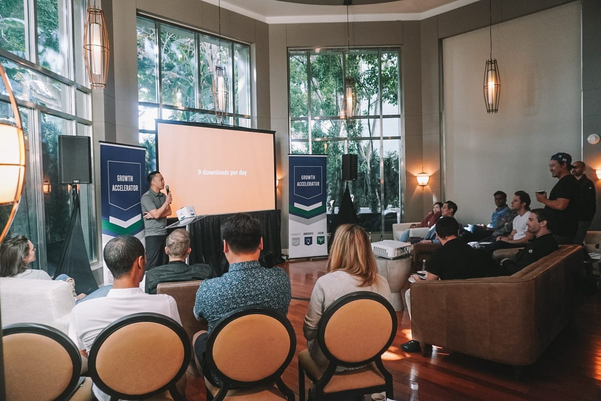A speaker with a microphone before a screen reading 9 downloads per day, between two Growth Accelerator banners