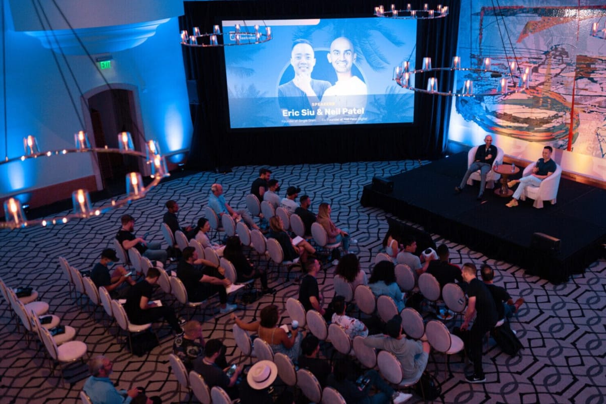 Audience in a patterned-carpet ballroom facing a stage under a screen reading Eric Siu & Neil Patel