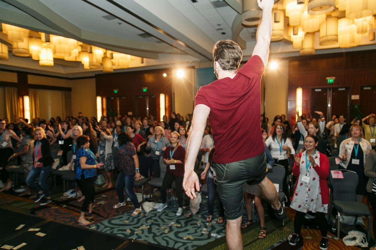 A speaker in a red T-shirt leaping with one arm raised in front of a standing crowd wearing lanyards