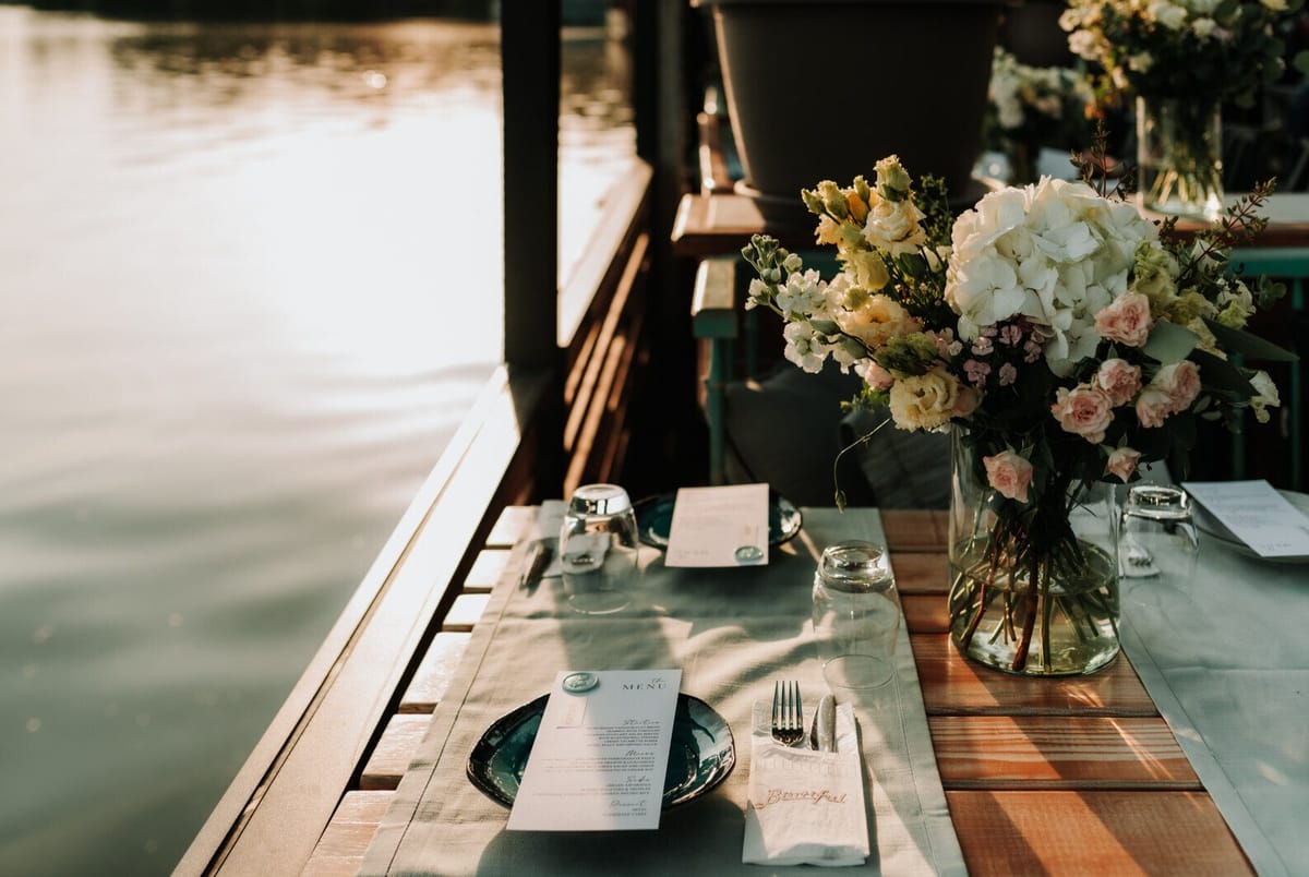 Table on a waterside deck set with a sage runner, menu cards and a vase of hydrangeas and roses