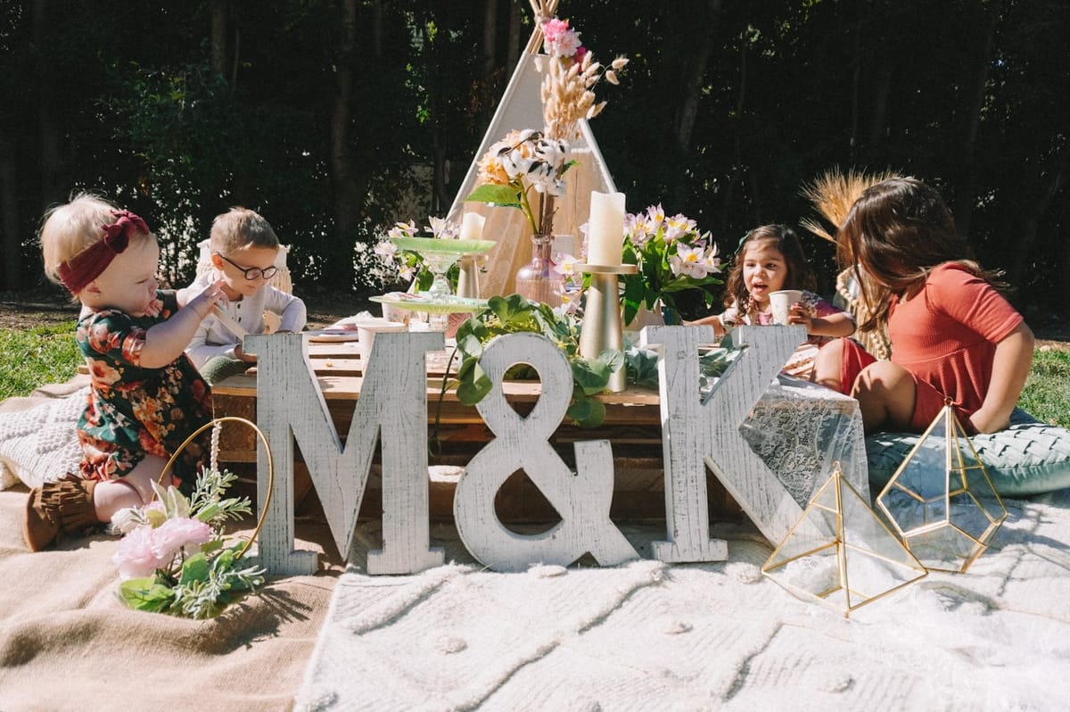 Children playing at a low pallet table behind whitewashed M&K letters, with a white teepee and flowers behind