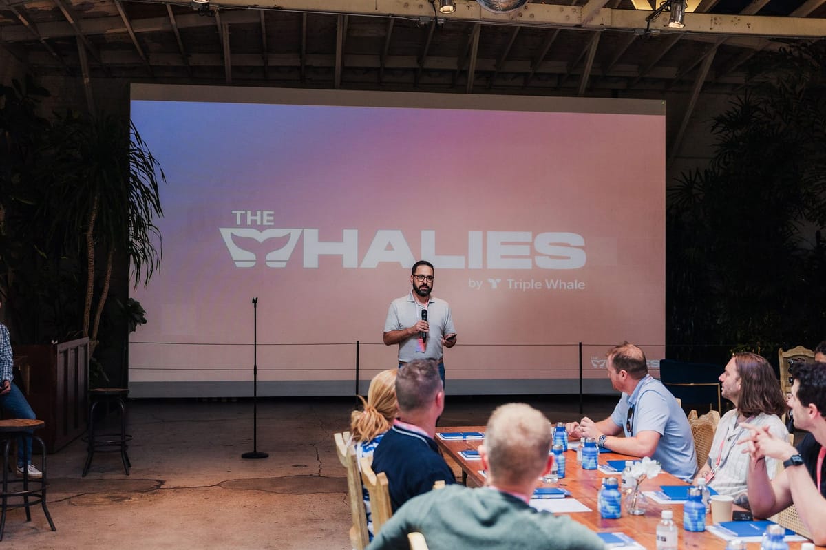 Speaker with a microphone in front of a screen reading The Whalies by Triple Whale, guests at a long table