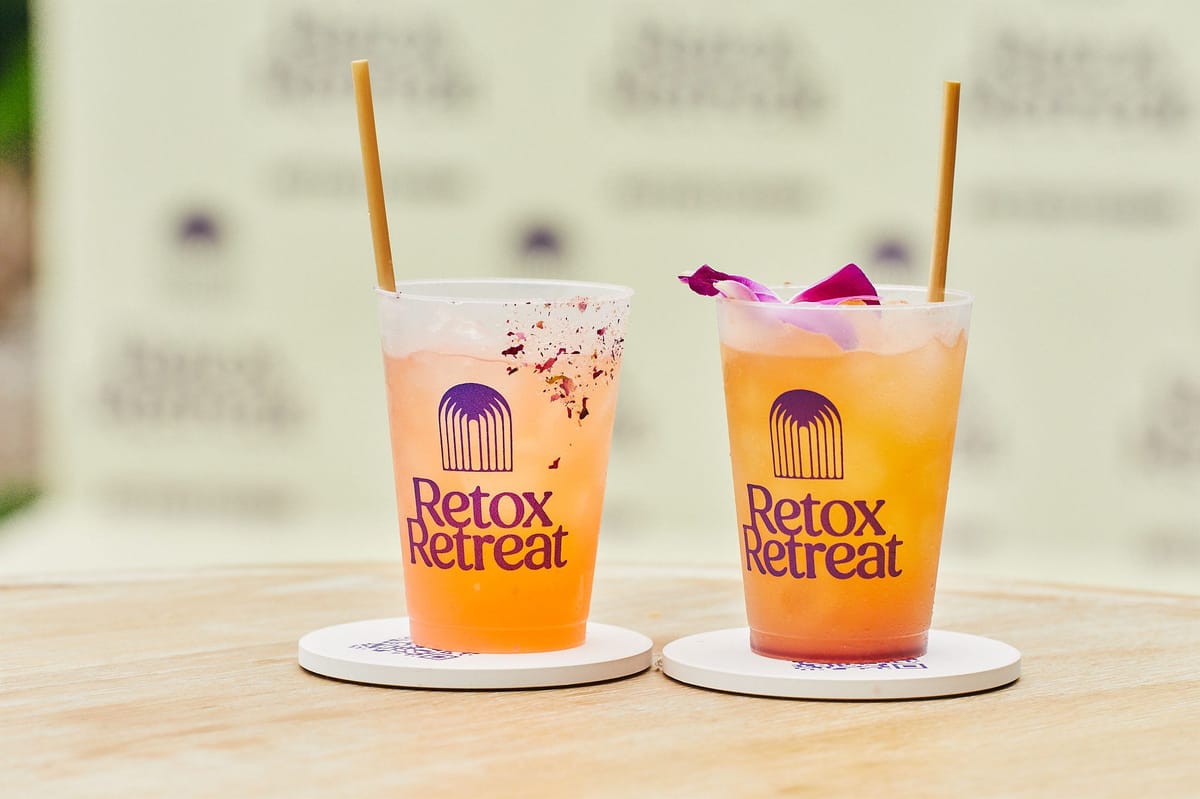 Two orange drinks in clear Retox Retreat cups with gold straws on white coasters, a logo backdrop blurred behind