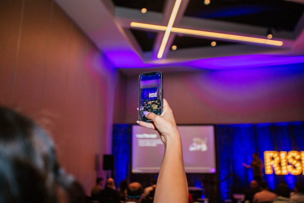 A guest holds up a phone toward the stage, where lit RISE marquee letters glow under purple light