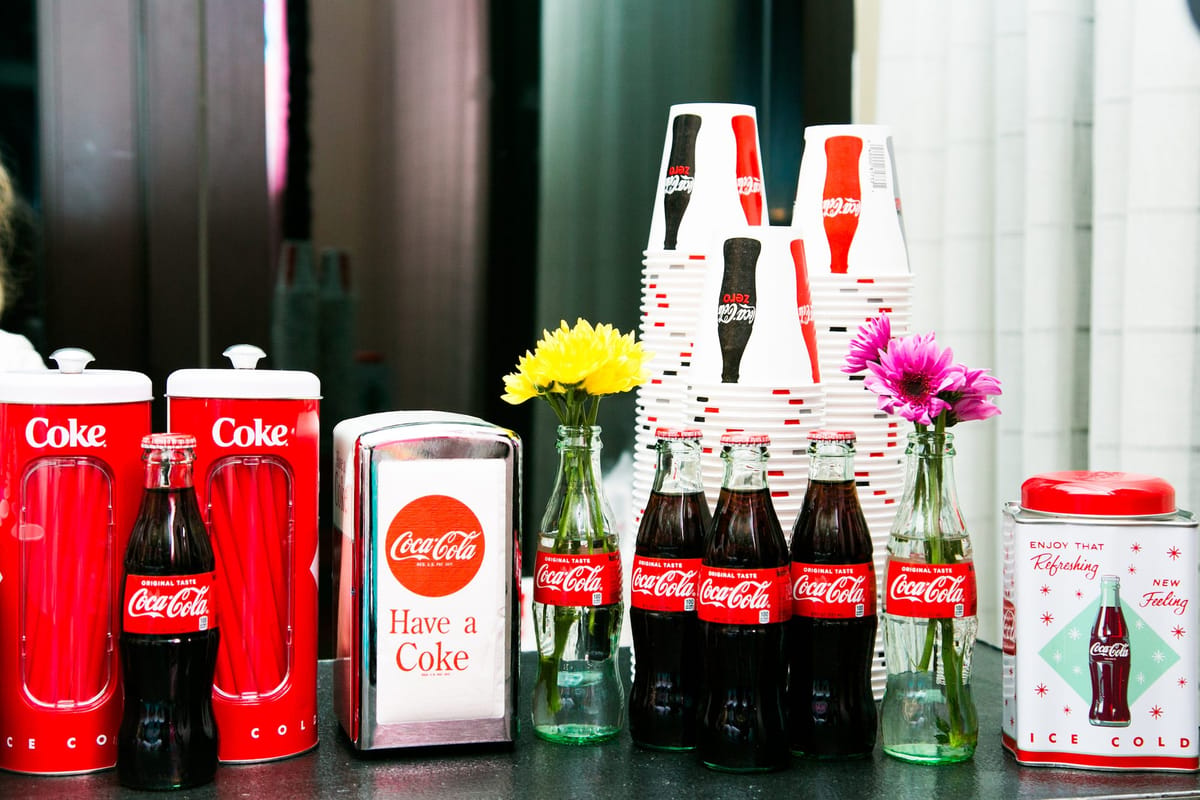 Coca-Cola bottles, red Coke straw tins, a Have a Coke napkin holder and stacked Coca-Cola cups with flowers