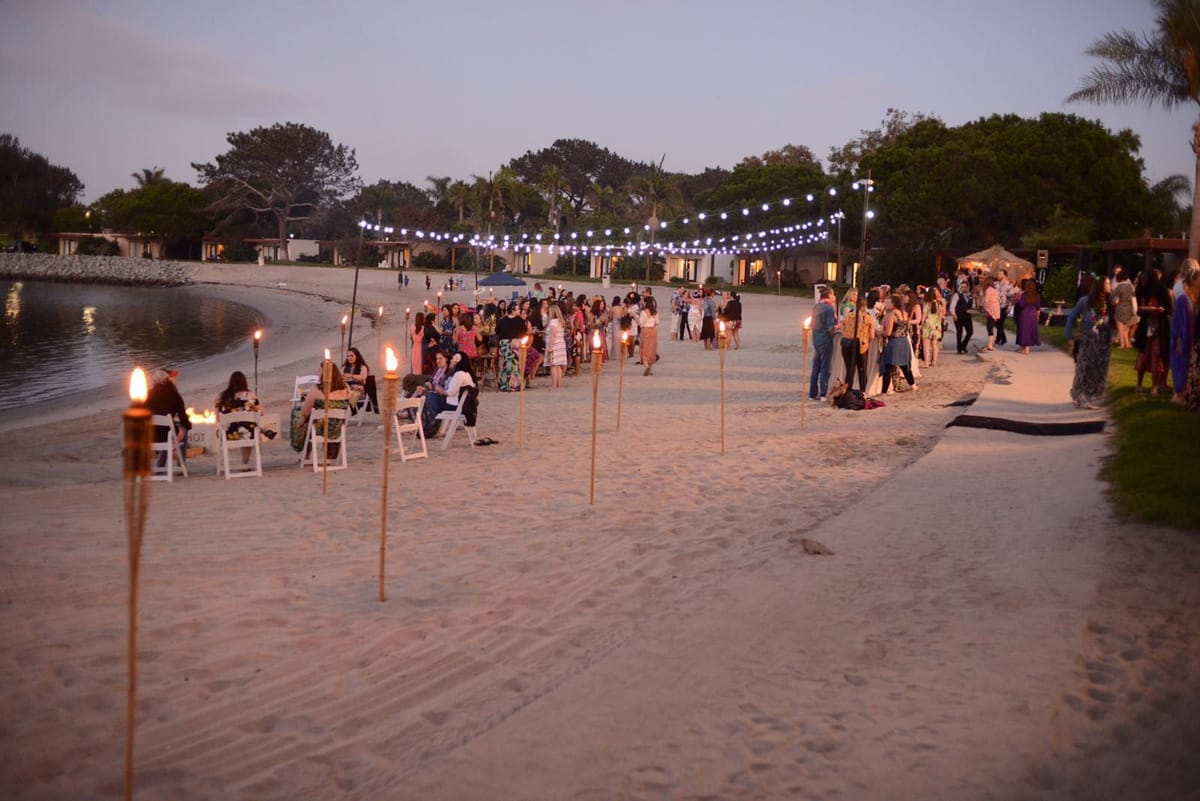 Guests at tables on a beach at dusk, tiki torches lining the sand and string lights along the shore