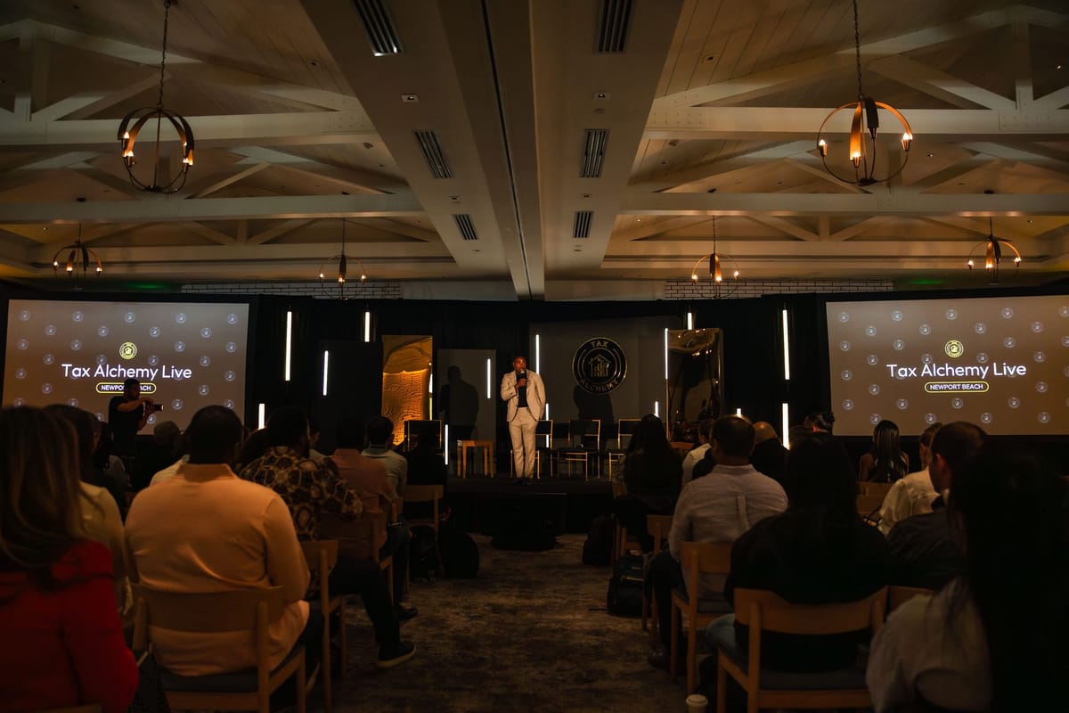 Audience facing a speaker in a white suit on a stage flanked by two Tax Alchemy Live screens