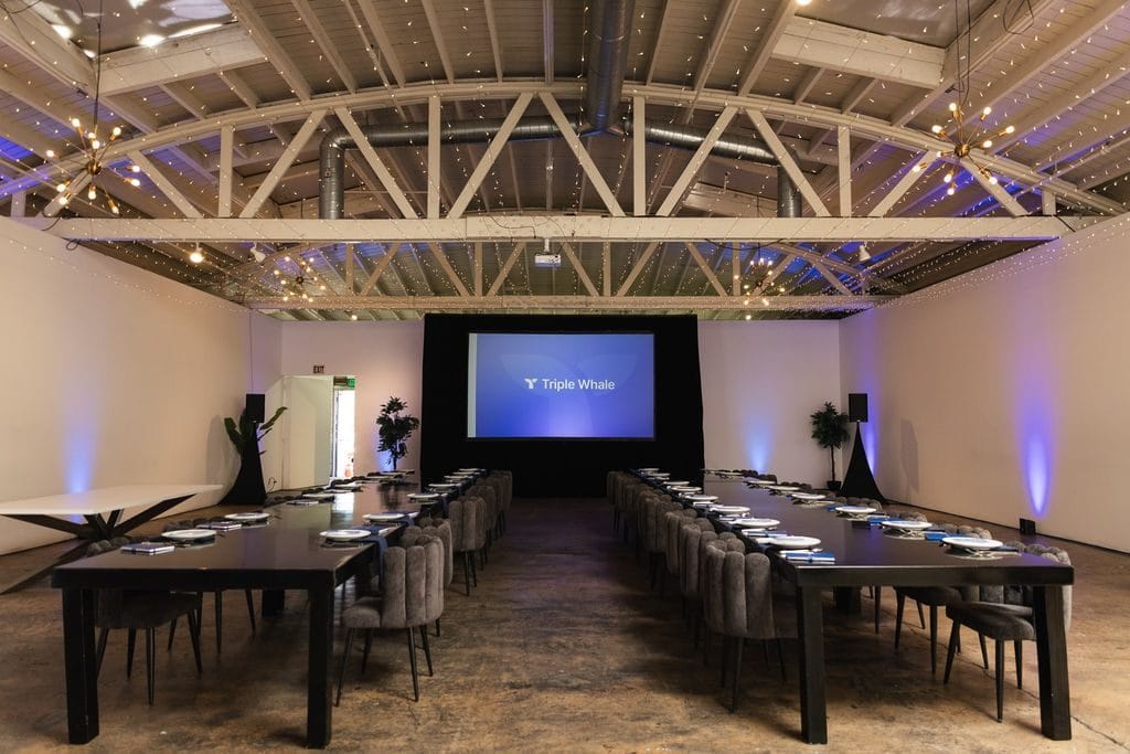 Empty boardroom with two long black tables, grey chairs and a Triple Whale screen under twinkle lights