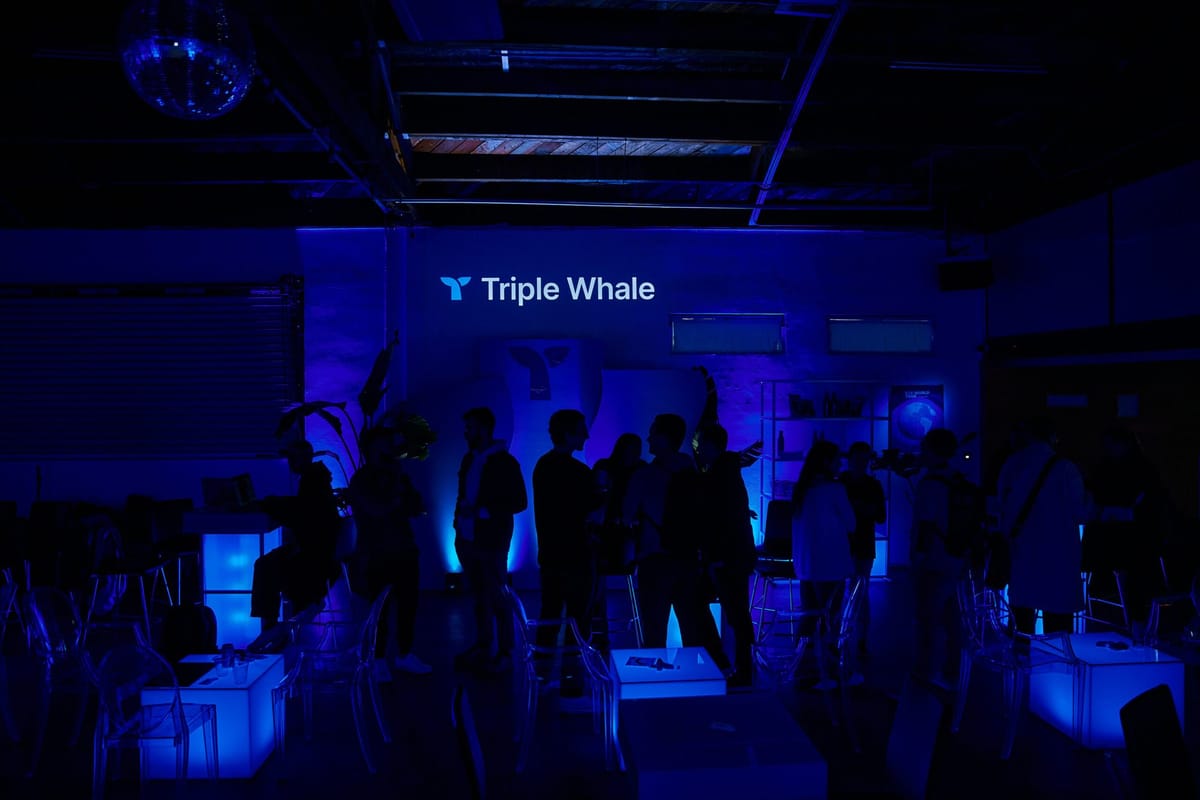 Guests in silhouette under Triple Whale lettering in a dim blue room with glowing cube tables and a disco ball