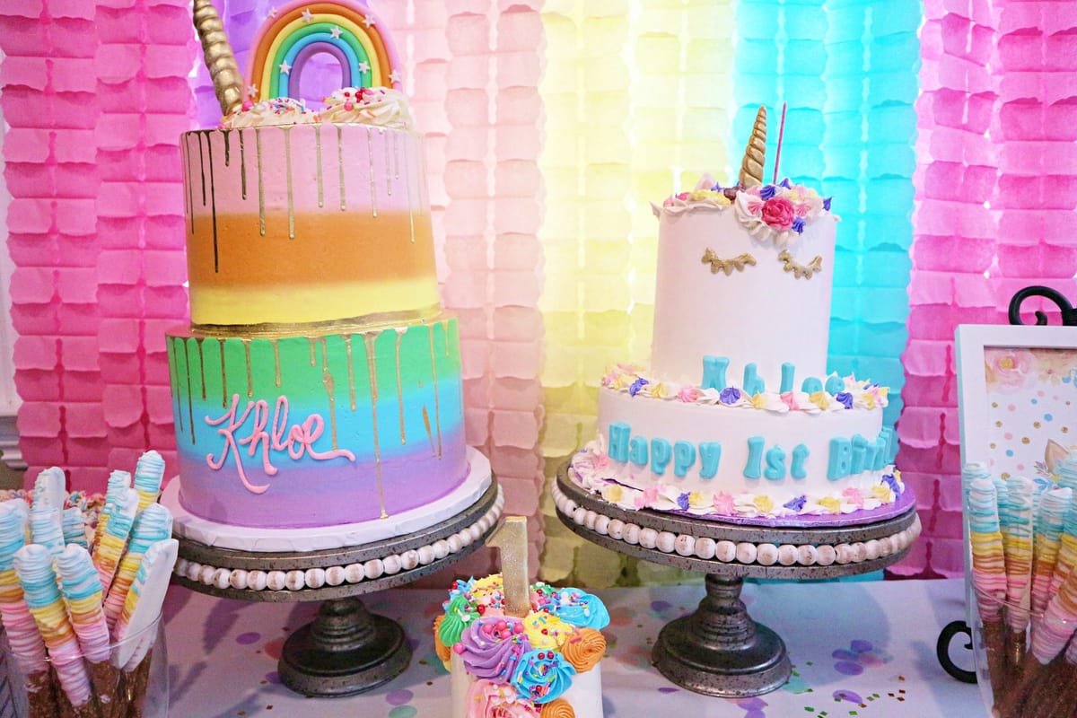 Rainbow-striped Khloe cake and a white unicorn cake reading Happy 1st Birthday in front of pastel paper garlands