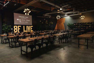 Long wooden communal tables facing illuminated 8F LIVE marquee letters and a sponsor screen