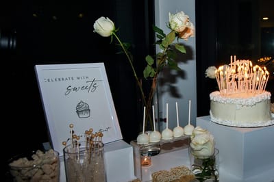 Sweets table at night with a Celebrate with sweets sign, white roses, white cake pops and a candlelit white cake