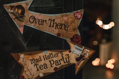 Wooden arrow signs reading Over There and Mad Hatter’s Tea Party with a Cheshire Cat illustration