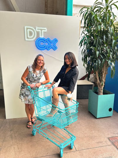 Samantha Pena and another woman with a turquoise shopping cart under a neon DTCx sign