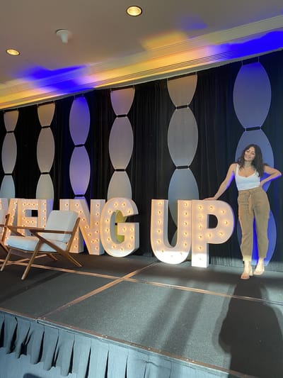 Samantha Pena posing on a stage beside giant marquee letters, with two lounge chairs to one side