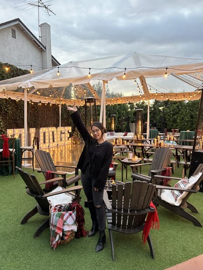Samantha Pena with one arm raised at an outdoor ski lodge party with Adirondack chairs, plaid blankets, string lights and LODGE marquee letters