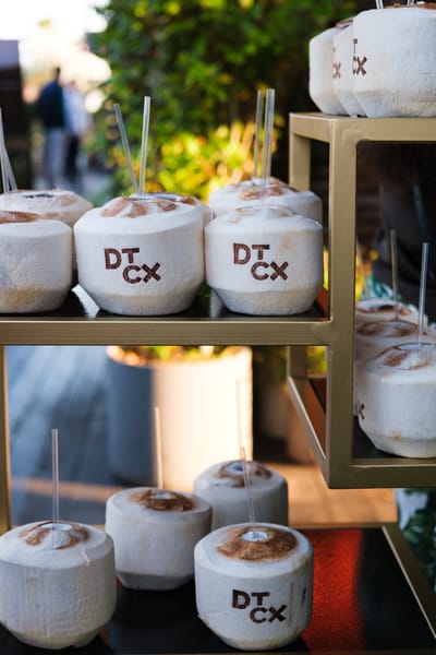 Fresh coconuts stamped with the DTCx logo, with straws, on gold shelving outdoors