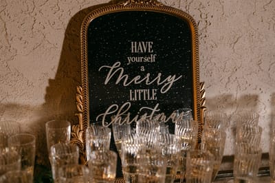 Gold framed mirror sign reading Have Yourself a Merry Little Christmas behind rows of clear glasses