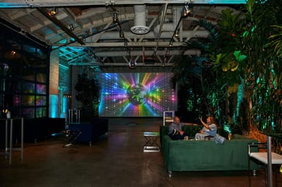 Warehouse party space with a rainbow disco ball video wall, green sofas and tall plants