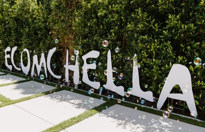Tall white ECOMCHELLA letters standing in front of a green hedge with soap bubbles floating past