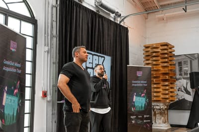 Two speakers in front of black curtains between GeekOut Miami Acquisition Tour banners in a white brick room