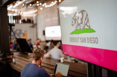 Screen showing a California bear and GEEKOUT SAN DIEGO above a pink band, with attendees on laptops behind