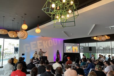 Audience watching a speaker in a bright room with GeekOut projected on the wall and a welcome slide on screen