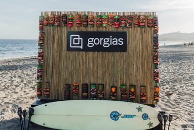 Bamboo wall trimmed with carved tiki masks and a Gorgias sign on a beach, with a surfboard and tiki torches