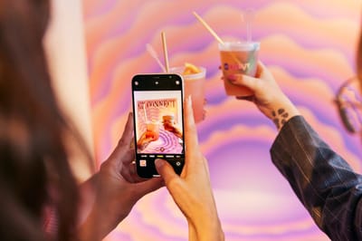 Hands toasting drinks, one in a Rebuy cup, while a phone photographs them against a CX Connect wave wall