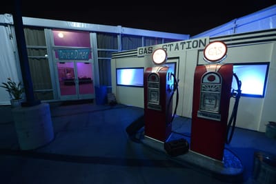 Two red vintage gas pumps with glowing GAS globes under a GAS STATION sign, beside Drive’n’Diner doors at night