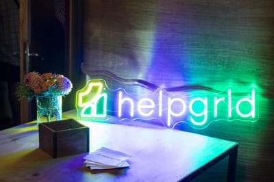 Green and blue HelpGrid neon sign on a table with a vase of dahlias, a wooden box and napkins