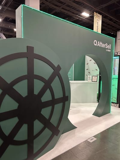 A bank vault door built at aisle scale into the green AfterSell by Rokt booth wall, with seating behind it