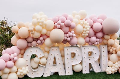 Oversized CAPRI marquee light-up letters set in a pink, peach and cream balloon wall on a lawn