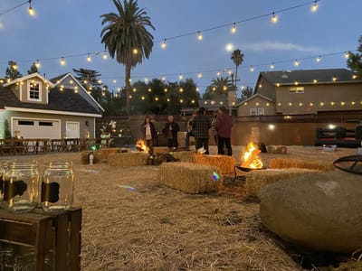 Straw-covered yard with hay bale seats and fire pits under string lights at dusk