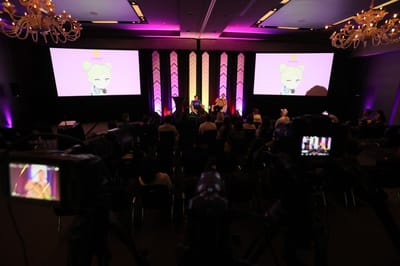 Audience facing two screens and purple-lit chevron pillars at Leveling Up Austin, with a camera filming