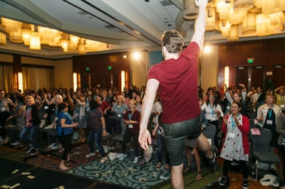 A speaker in a red T-shirt leaping with one arm raised in front of a standing crowd wearing lanyards