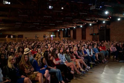 Packed audience seated in rows in a brick-walled hall under spotlights and exposed ceiling beams