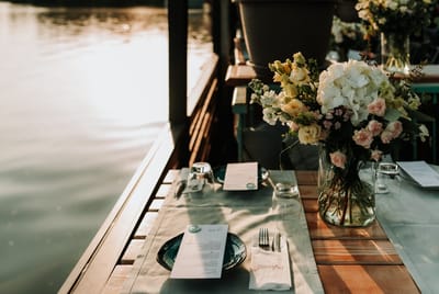 Table on a waterside deck set with a sage runner, menu cards and a vase of hydrangeas and roses