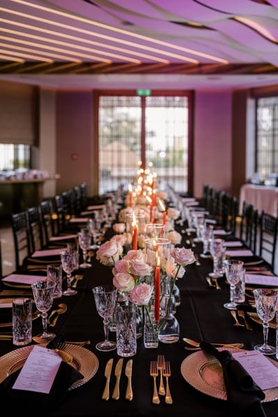 Long table set with gold chargers, menu cards, pink roses and orange taper candles in a pink-lit room