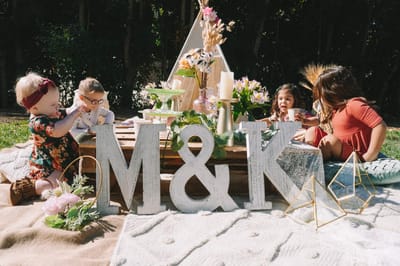Children playing at a low pallet table behind whitewashed M&K letters, with a white teepee and flowers behind