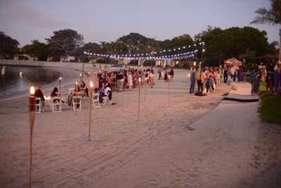 Guests at tables on a beach at dusk, tiki torches lining the sand and string lights along the shore