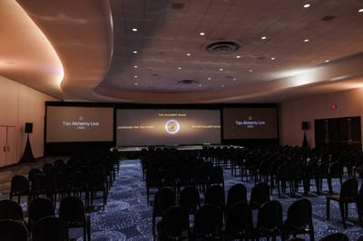 Dim conference room with rows of black chairs facing three screens under a curved, lit ceiling